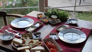 A dining table set with various cheeses, charcuterie, and bread, with a view of a pool and outdoor seating in the background. - Alaçatı Otel Rebetiko (Cesme)
