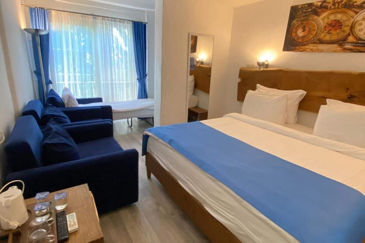 A hotel room with a large bed, a sofa, a small table with toiletries, a window with curtains, and a wall clock.