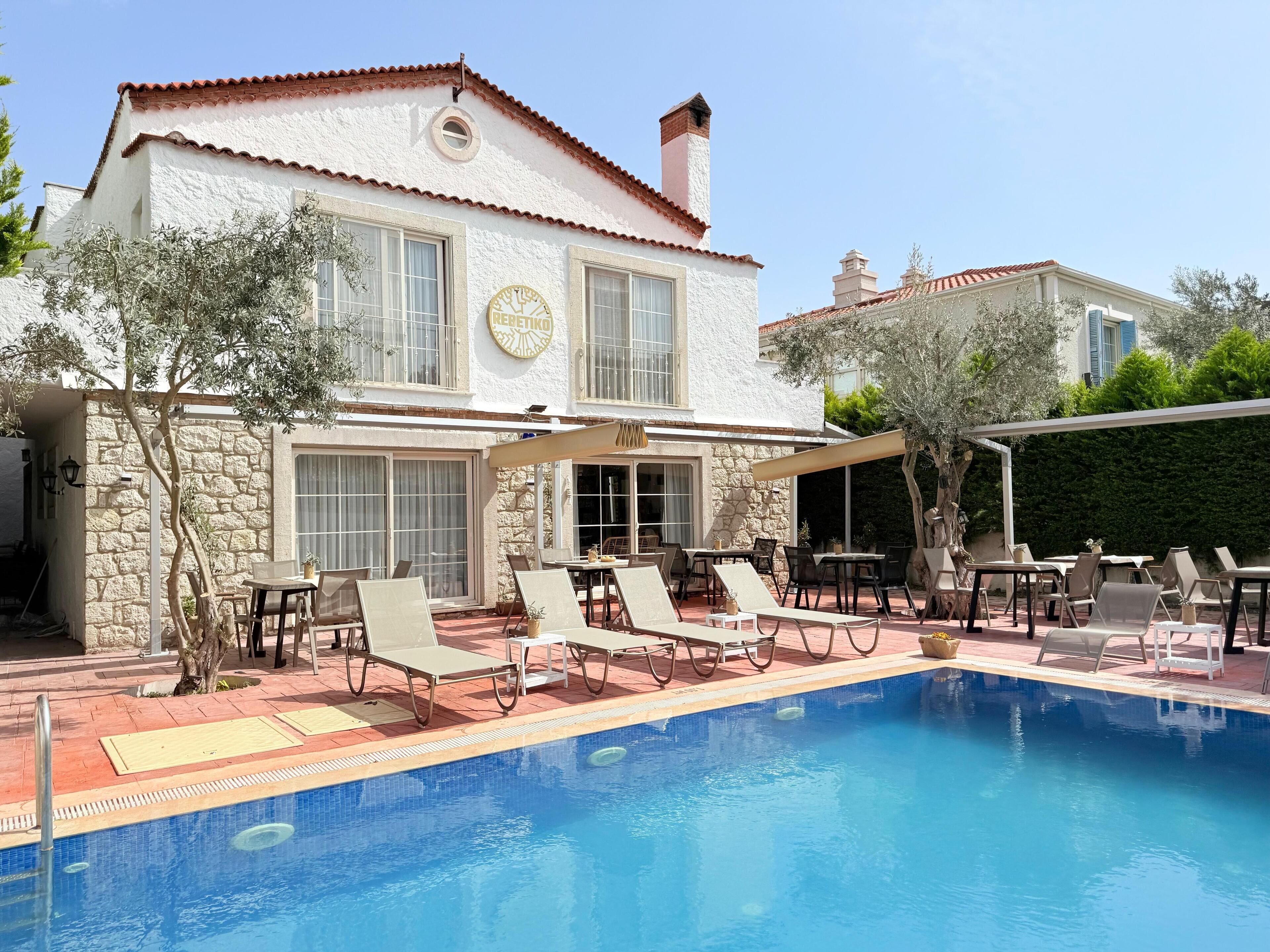 A poolside area with lounge chairs and tables, a stone wall, and a building with a tiled roof.