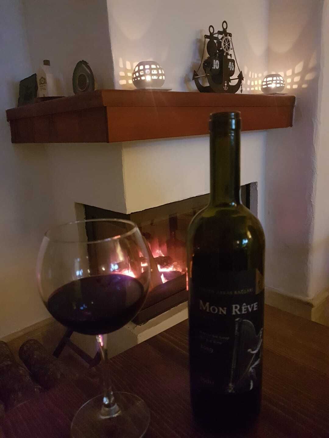 Una botella de vino Mon Rêve y una copa de vino tinto sobre una mesa frente a una chimenea encendida.