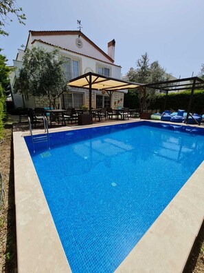 A rectangular swimming pool with a tiled edge, surrounded by a patio area with outdoor furniture and a covered seating area. - Alaçatı Otel Rebetiko (Cesme)