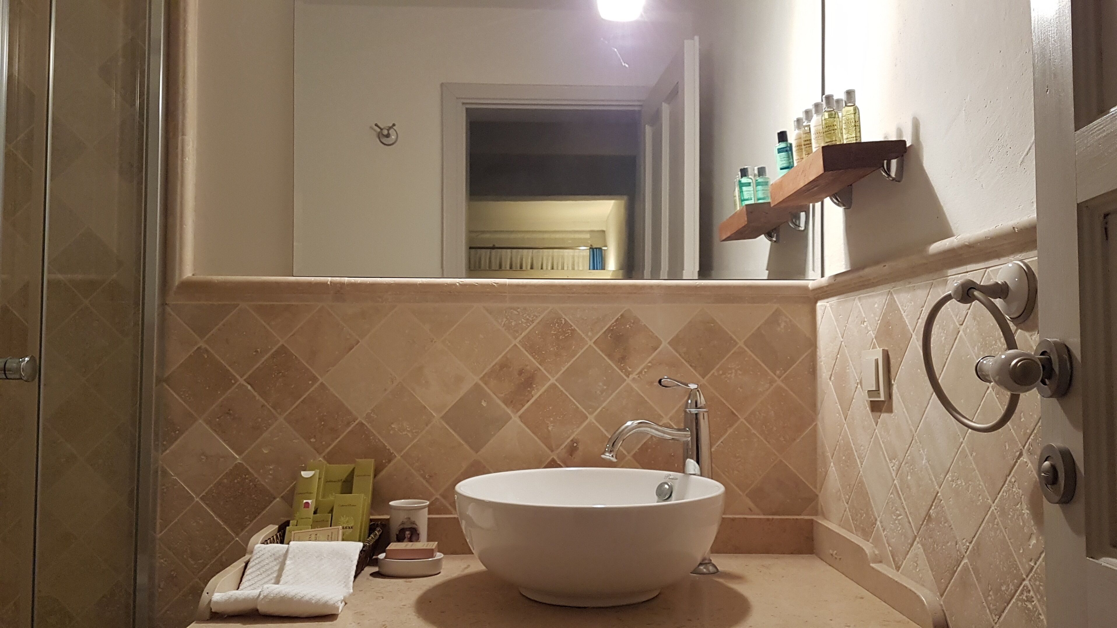A bathroom with a large sink, a towel, toiletries, and a mirror reflecting a window and a shelf with bottles.
