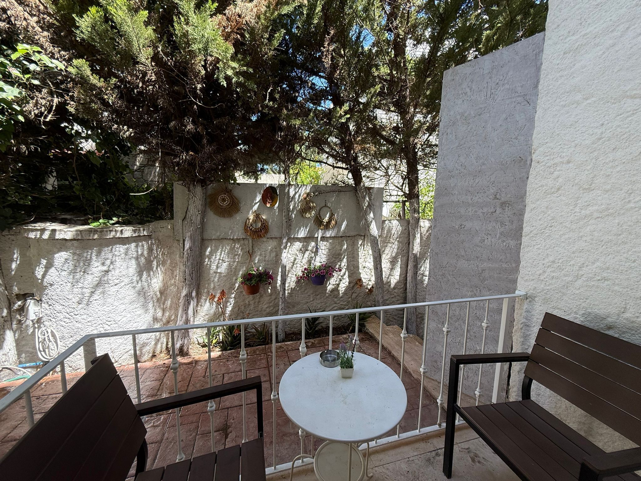 A balcony with a white table and two wooden chairs, surrounded by a white wall with decorative wall hangings.
