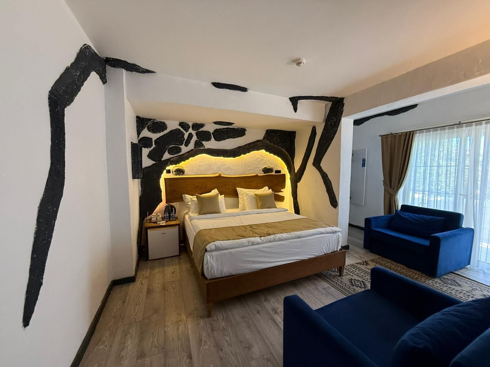 Habitació d'hotel amb un llit, una tauleta de nit, un butàc blau i un gran finestral amb cortines.