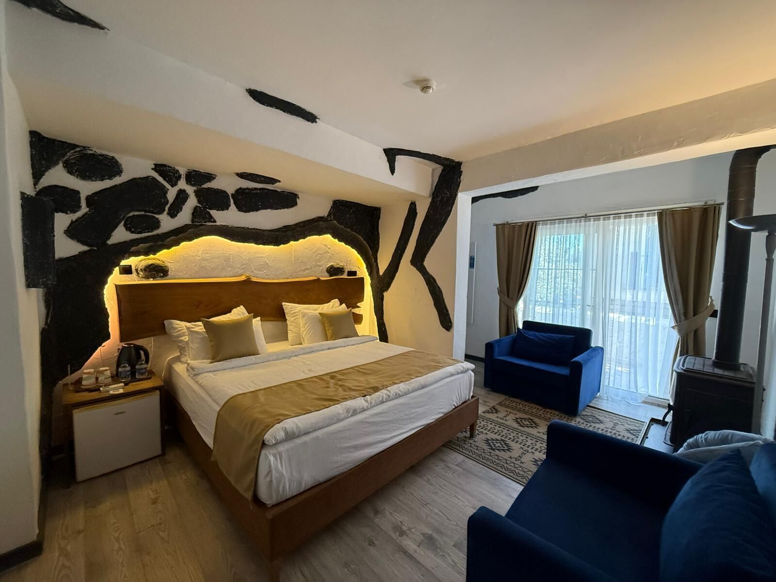 Ein Hotelzimmer mit Bett, Nachttisch, blauem Sessel, Fenster mit Vorhängen und Kamin.