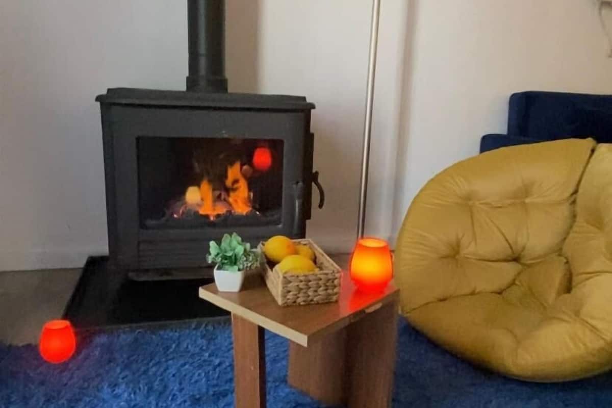 A cozy living room with a wood-burning stove, a small wooden table with a basket of lemons, a yellow armchair, and a tall floor lamp.
