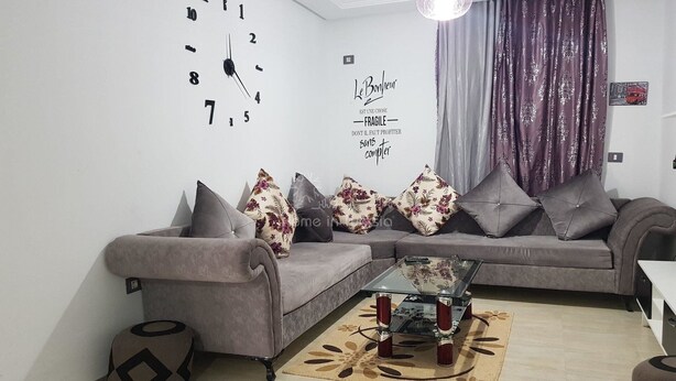 Ein modernes Wohnzimmer mit einem grauen Sofa, dekorativen Kissen, einem Glastisch und einer Wanduhr.