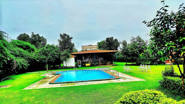 A swimming pool with a surrounding lawn and a covered seating area. - Hills Nature Resort (Gurugram)