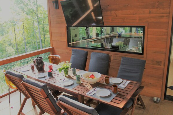 A dining area with a wooden table set for four, surrounded by chairs, a TV mounted on the wall, and a view of the outdoors. - Modern Mountain Retreat in Serene Lake Community (Saluda)