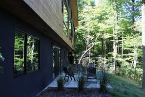 Una casa moderna con ampie finestre, un patio con mobili da esterno e uno sfondo naturale con alberi.