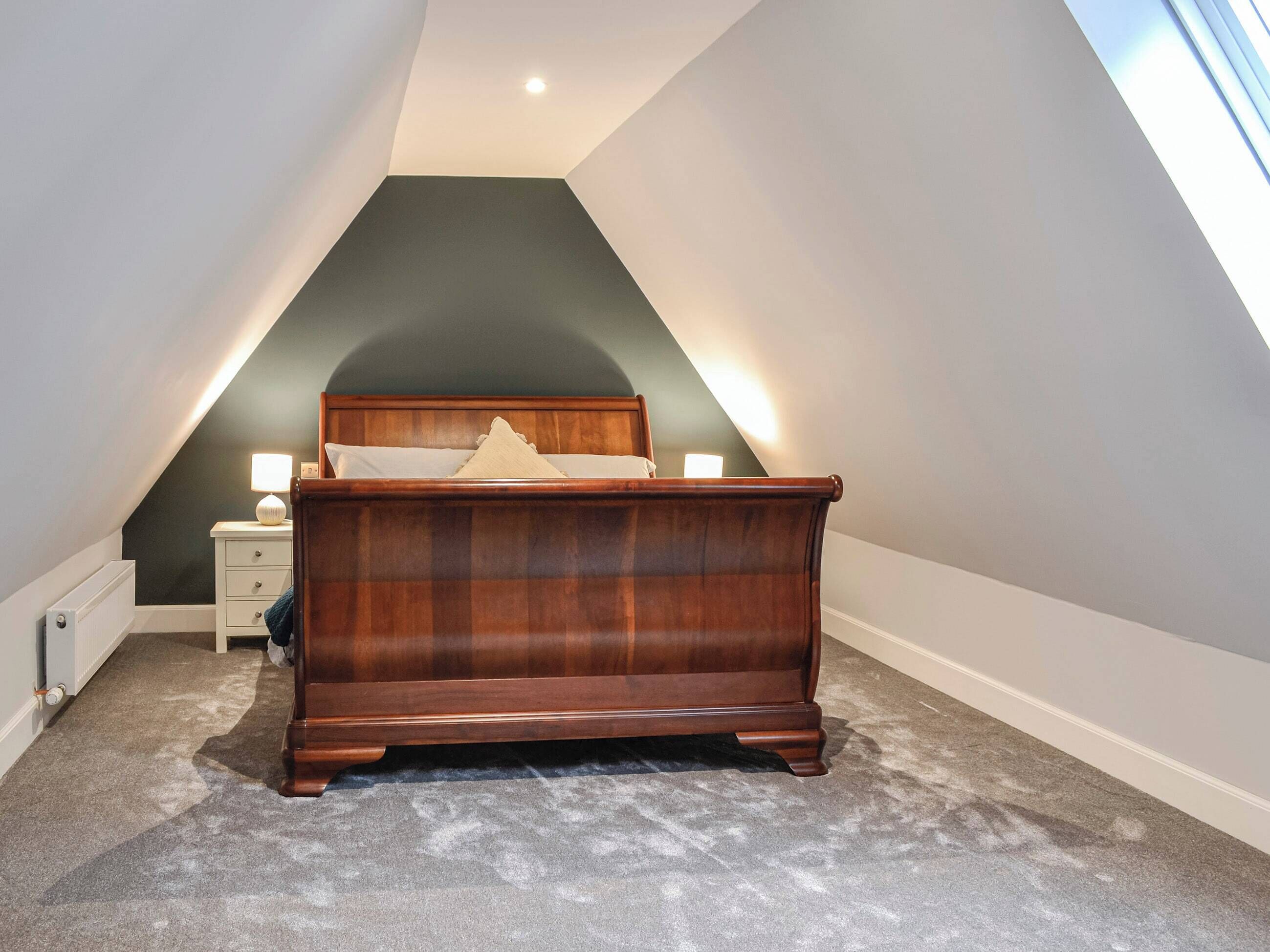 A bedroom with a sloped ceiling, a wooden bed, a nightstand, and a lamp.