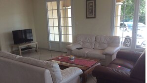 Ein Wohnzimmer mit einem beigefarbenen Sofa, einem braunen Sessel und einem Couchtisch. Ein Fernseher steht auf einem Schrank und ein Fenster bietet Blick auf parkende Autos vor dem Haus.