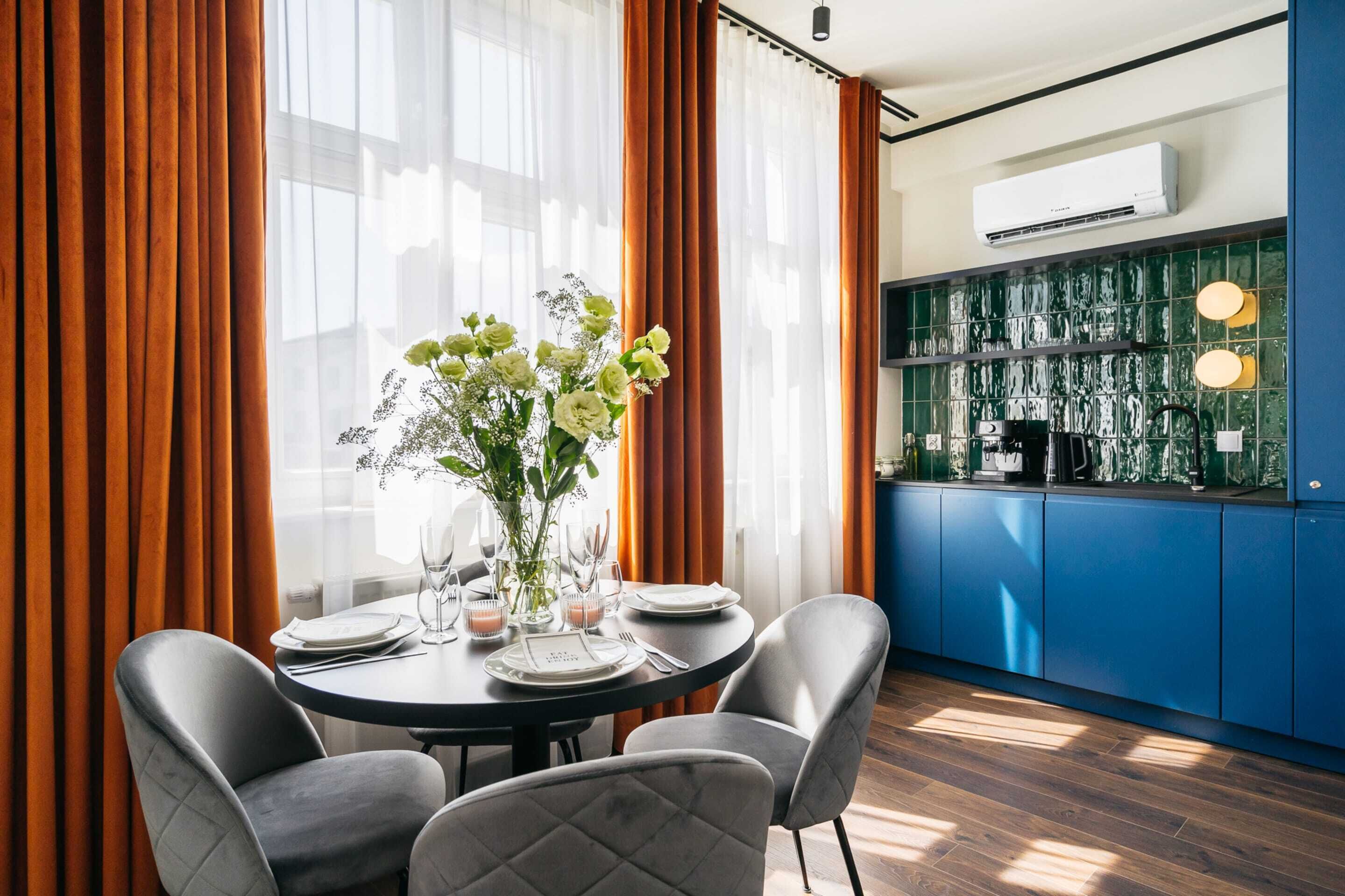 A dining area with a round table set for four, surrounded by grey upholstered chairs. A vase with white flowers is on the table, and a kitchen with blue cabinets and a green backsplash is visible in the background.