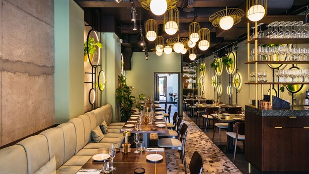A restaurant with a long communal dining table, cushioned seating, and a bar area with shelves of glasses. - Estery12 (Kraków)