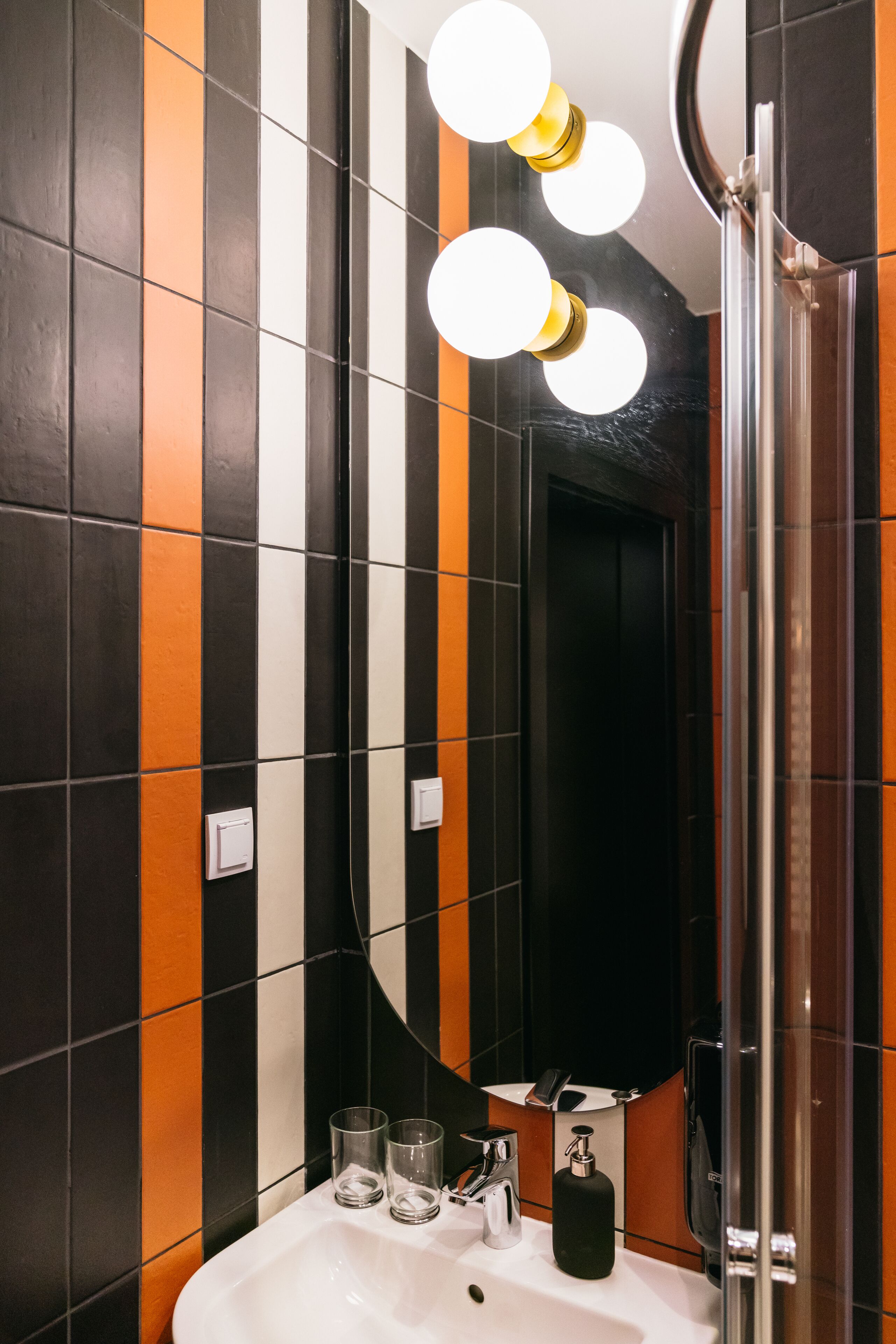 Une salle de bain avec des carreaux noirs et orange, un lavabo blanc, deux verres, un distributeur de savon et un miroir avec éclairage.
