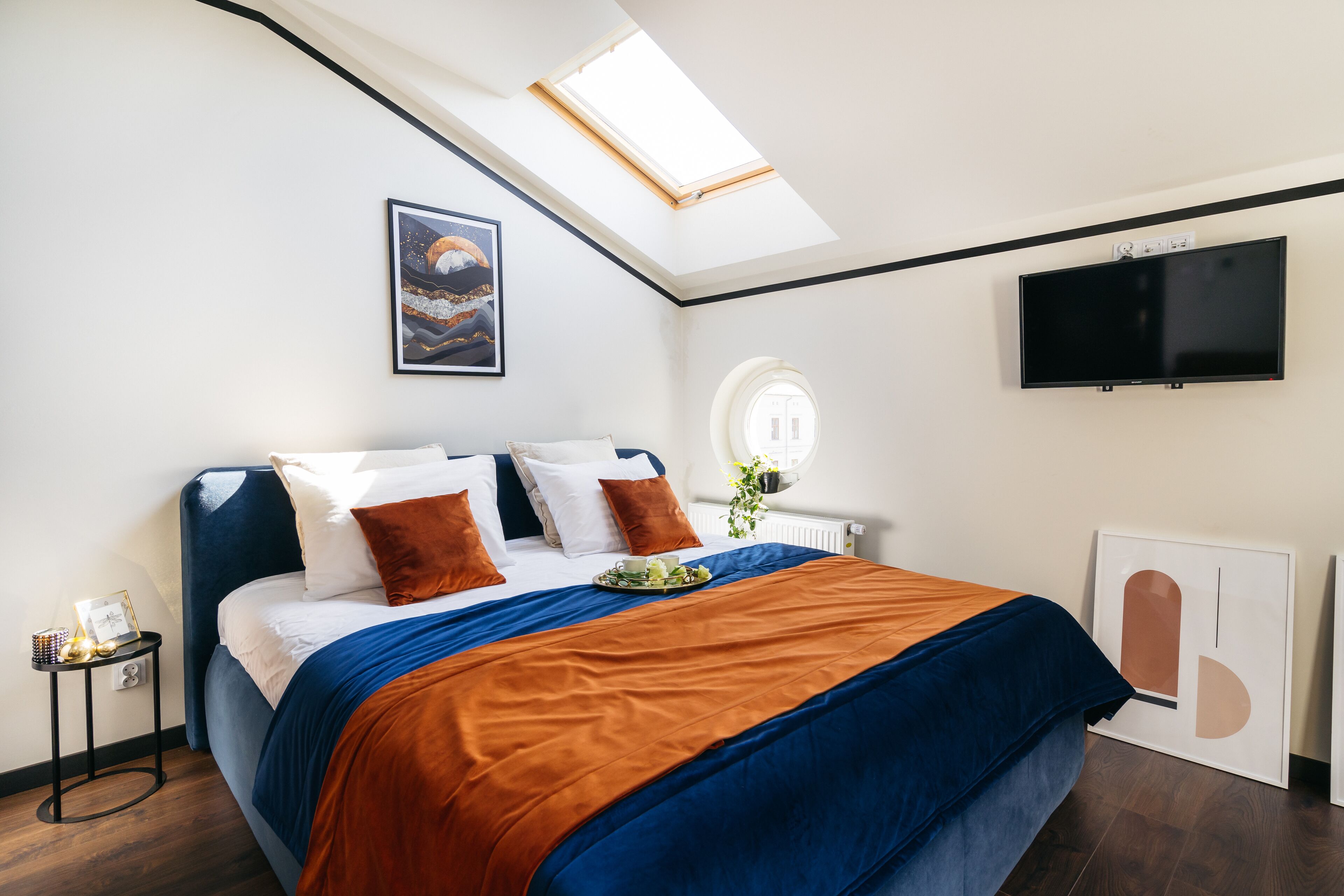 Quarto moderno com claraboia, cama com roupa de cama azul e laranja, televisão de tela plana e uma obra de arte emoldurada na parede.