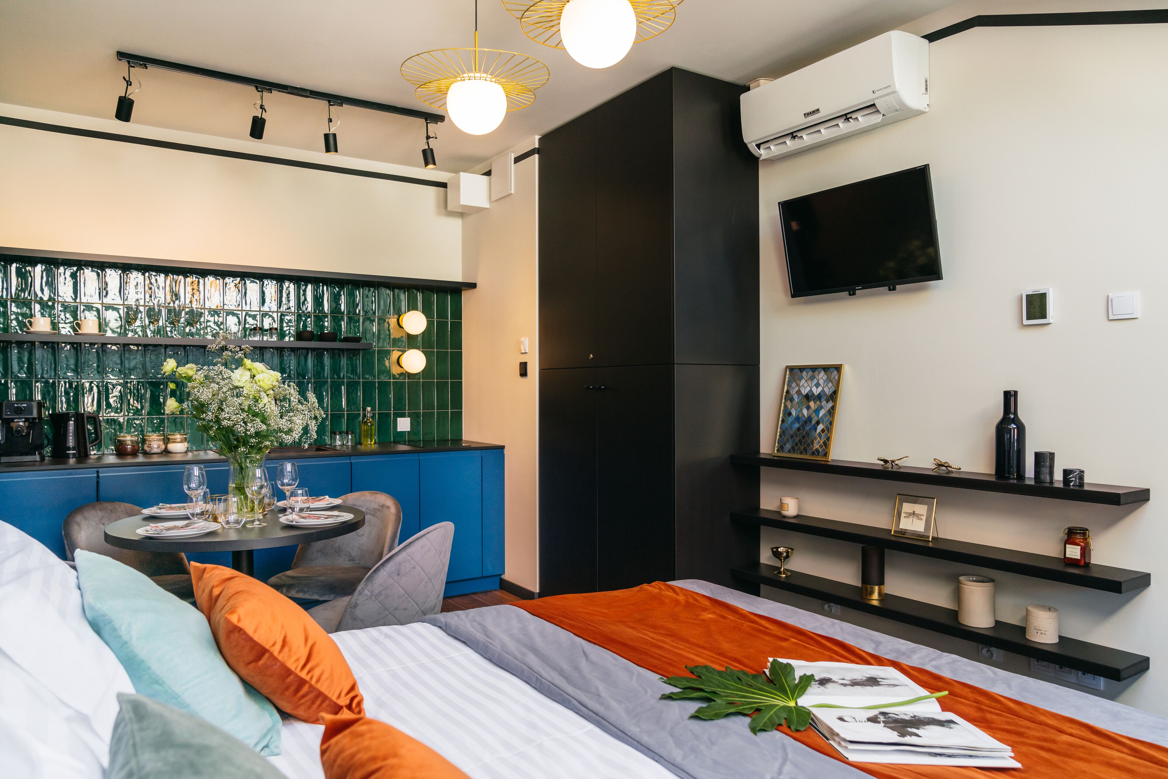 A modern hotel room with a bed, a dining table with chairs, a TV mounted on the wall, and a green-tiled backsplash.
