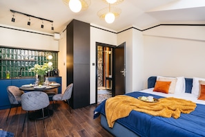 Habitació d'hotel moderna amb un llit, zona de dinar amb taula i cadires, i vista per una finestra amb marc verd.