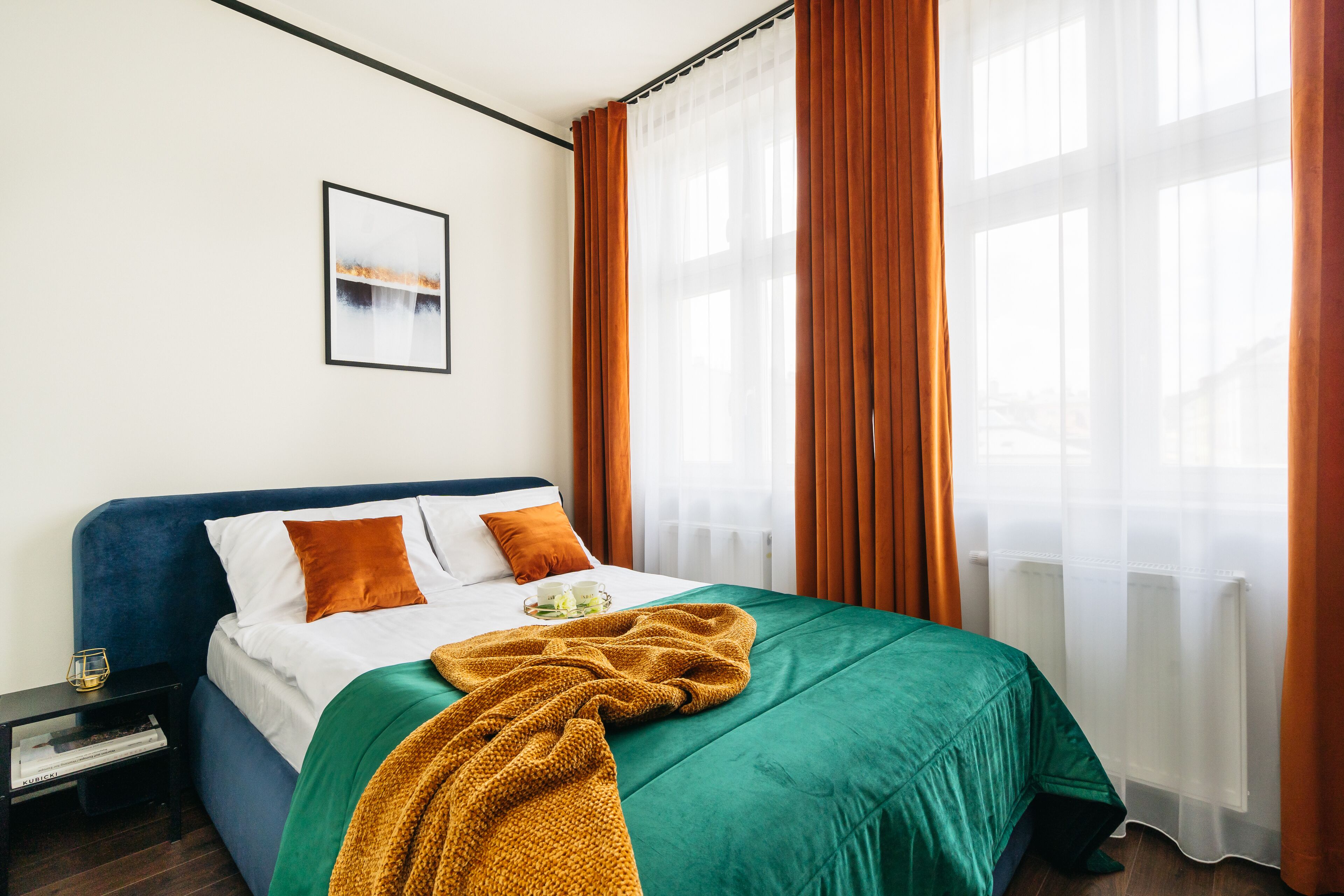 A neatly made bed with a green and white duvet, a mustard-colored throw blanket, and two orange pillows. A framed picture hangs on the wall above the bed.