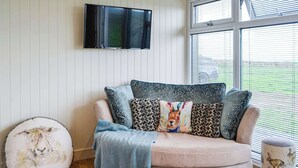 A living room with a beige sofa, patterned cushions, a round pouf with a sheep design, a blue throw blanket, and a television mounted on the wall.