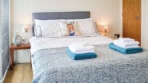 A neatly made bed with a patterned bedspread, two folded towels, decorative pillows, bedside lamps, and a wooden bedside table with a clock.