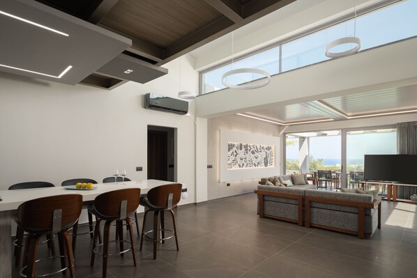 A spacious modern living area with a dining table, bar stools, a sofa, and a large flat-screen TV.