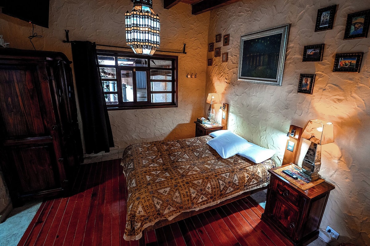 Ein Schlafzimmer mit Bett, Holzschrank, einer Chandelierleuchtre, einem Fenster und einer Wand mit gerahmten Bildern.