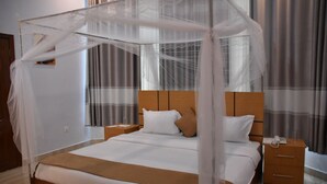 A hotel room with a four-poster bed, a wooden headboard, a bedside table with a lamp, and a nightstand with a phone. - Chimpanzee Lodge (Bushekeri)
