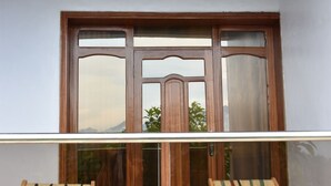 A balcony with two wooden lounge chairs, a small table, and a large window with a view of greenery and mountains. - Chimpanzee Lodge (Bushekeri)