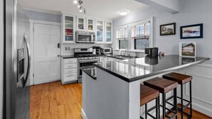 Una cocina moderna con armarios blancos, electrodomésticos de acero inoxidable, una isla grande con taburetes y una ventana con persianas.