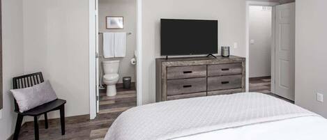 A bedroom with a bed, a TV, a dresser, a chair, and a bathroom visible through an open door.
