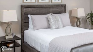 Una cama bien hecha con cabecera de madera, dos mesitas de noche con lámparas, y tres obras de arte abstracto enmarcadas en la pared.