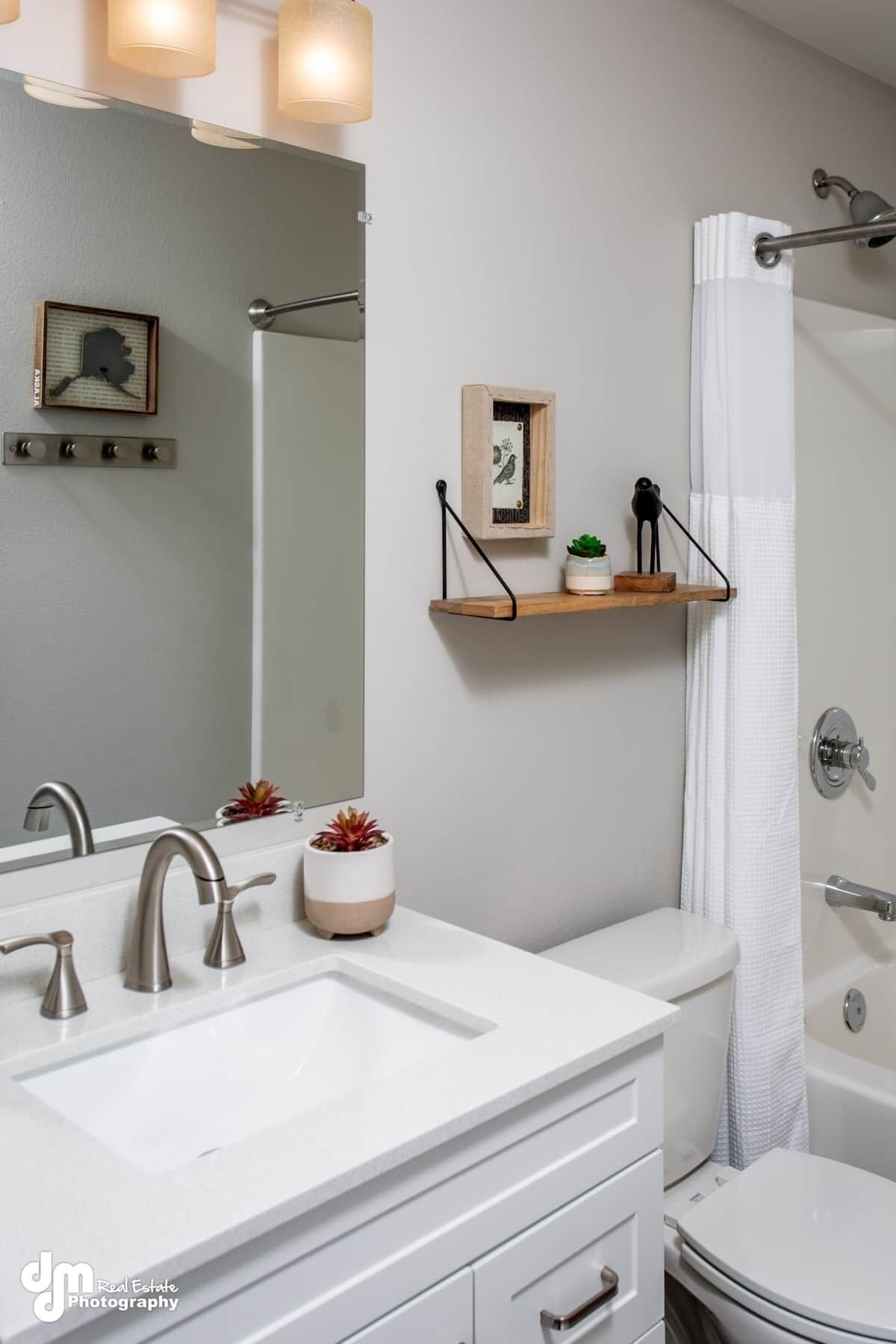 A modern bathroom with a white sink, two faucets, a mirror, and a shower curtain. There are two wall-mounted shelves with decorative items and a small plant.