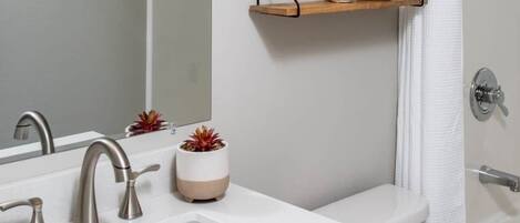 A modern bathroom with a white sink, two faucets, a mirror, and a shower curtain. There are two wall-mounted shelves with decorative items and a small plant.