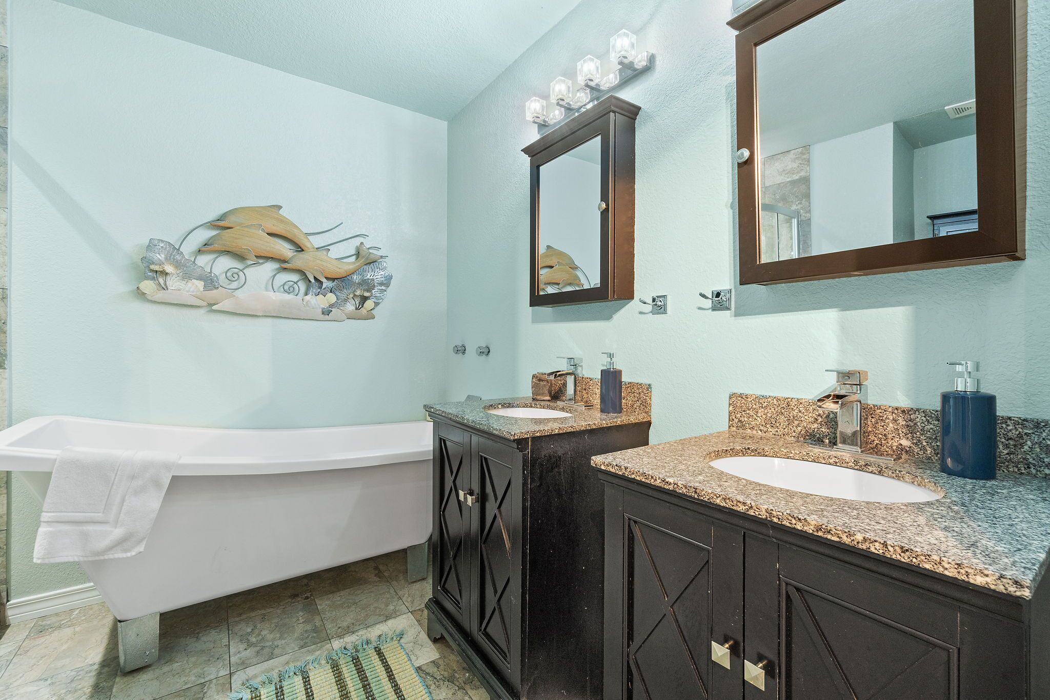 A bathroom with a white bathtub, two sinks with granite countertops, a large mirror, and a wall-mounted light fixture.