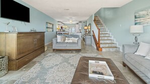 A spacious living room with a large flat-screen TV, a sofa, a coffee table, and a staircase leading to the upper floor.
