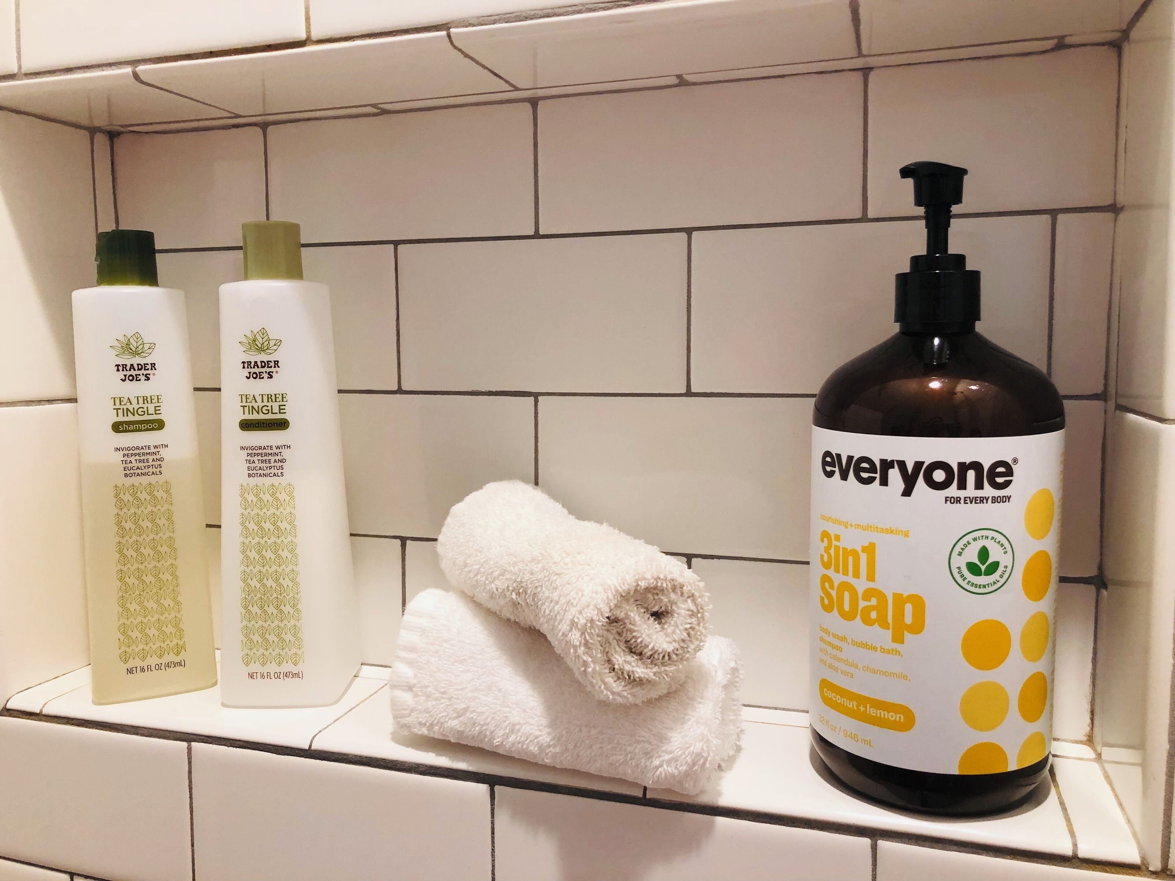 A bathroom shelf with three bottles of Trader Joe's Tea Tree Tingle shampoo and conditioner, and a bottle of Everyone 3in1 Soap.