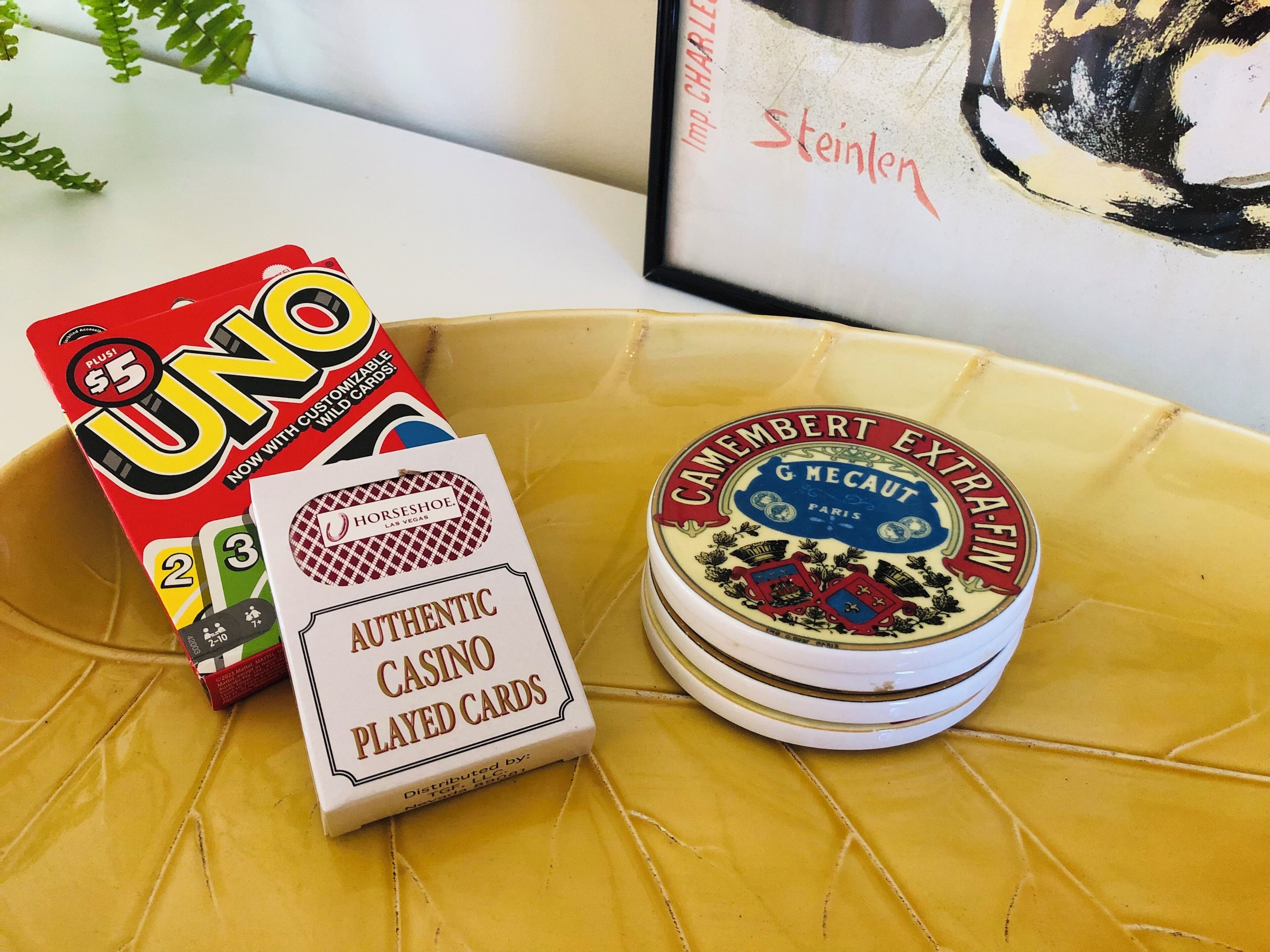 A box of UNO cards, a tin of Camembert cheese, and a framed painting on a yellow surface.