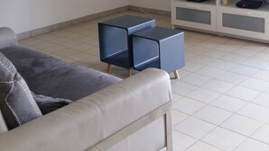 Un salon avec un canapé, deux poufs bleus, un téléviseur posé sur un meuble blanc et un escalier doté d’une rampe.