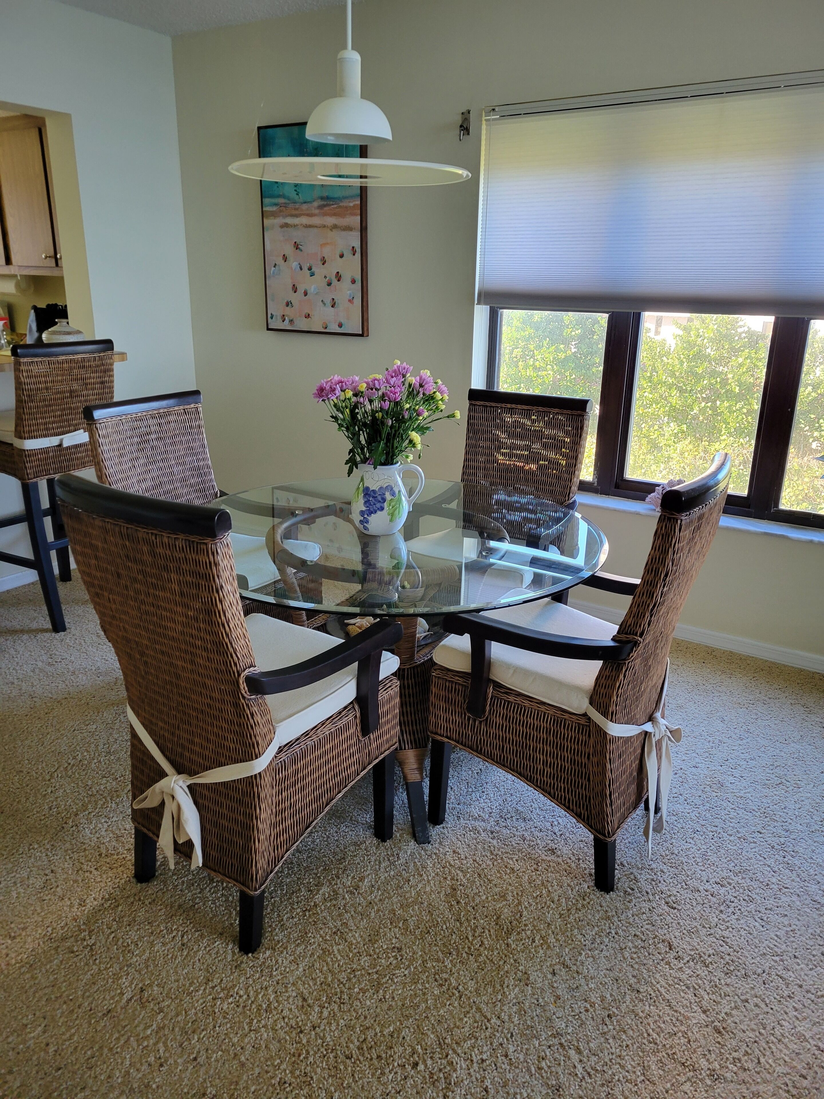 A dining area with a glass table surrounded by wicker chairs, a vase of flowers, and a painting on the wall.