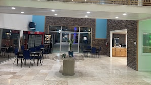 A spacious lobby with a high ceiling, a central round table with chairs, a vending machine, and a seating area with blue chairs. - B&J Hotel and Apartment (Pecos)