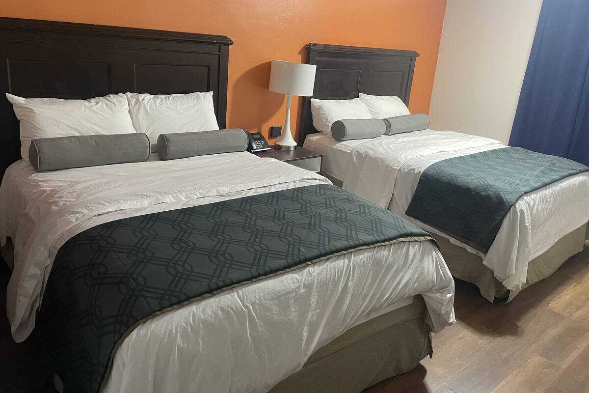 Une chambre d’hôtel avec deux lits, chacun doté de draps blancs et d’une tête de lit grise. Il y a une table de chevet avec une lampe et un téléphone. Les murs de la chambre sont de couleur orange et le sol est en bois.