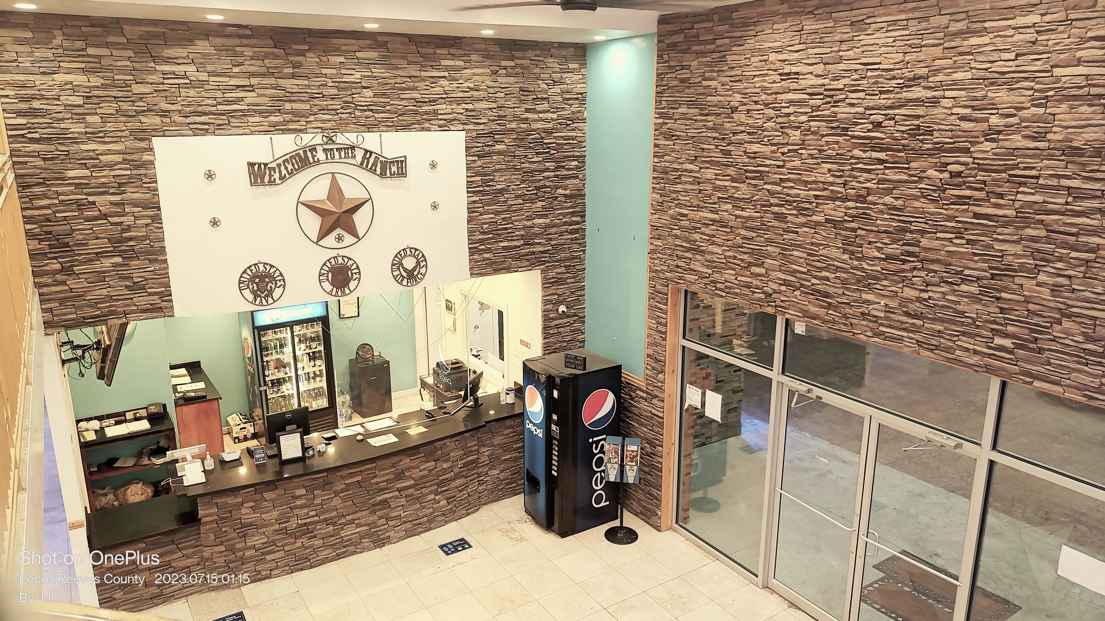 A reception area with a stone wall backdrop, a 'Welcome to the Ranch' sign, a Pepsi refrigerator, and a counter with various items.