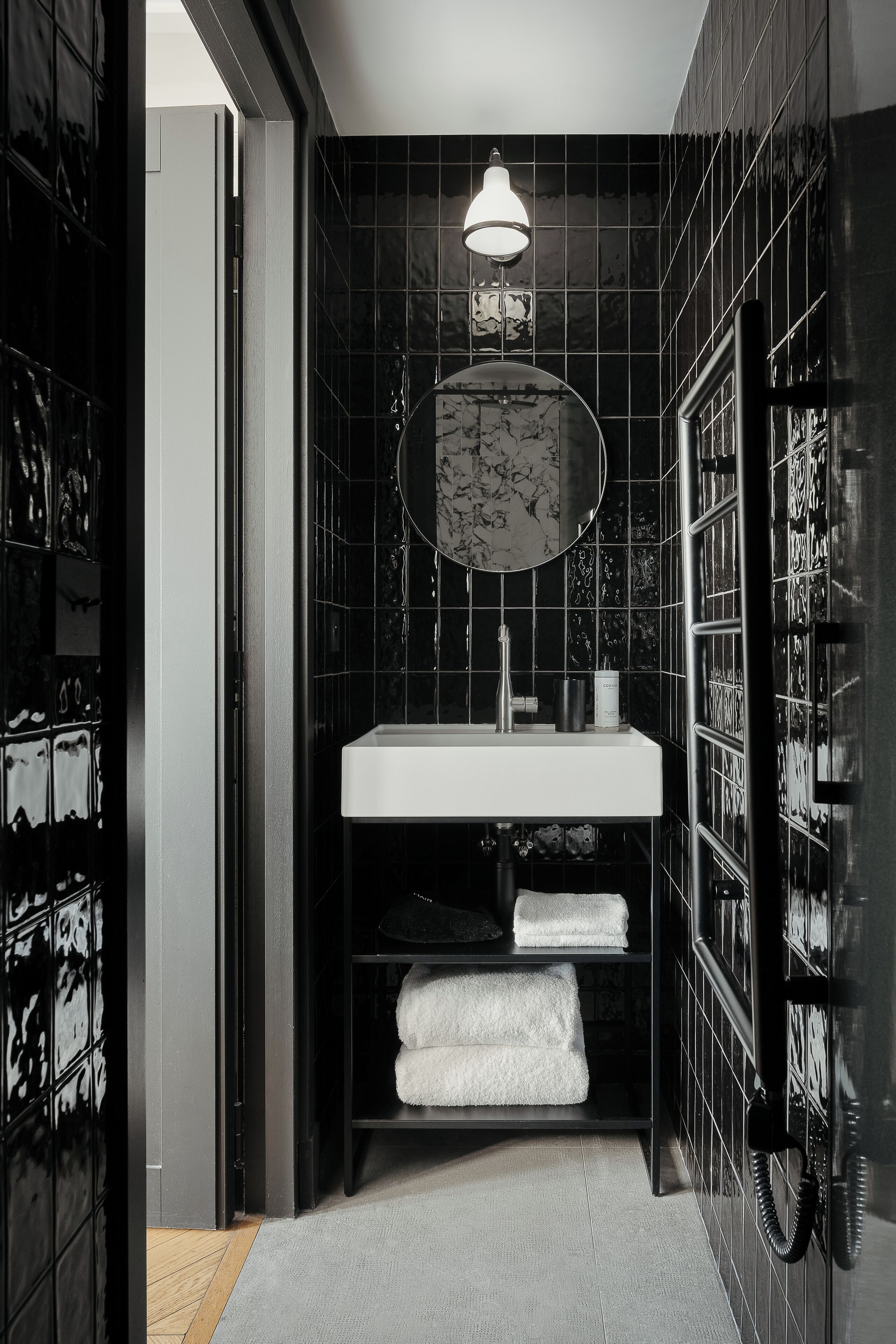 A modern bathroom with a white sink, black tiled walls, a round mirror, and a towel rack with folded towels.