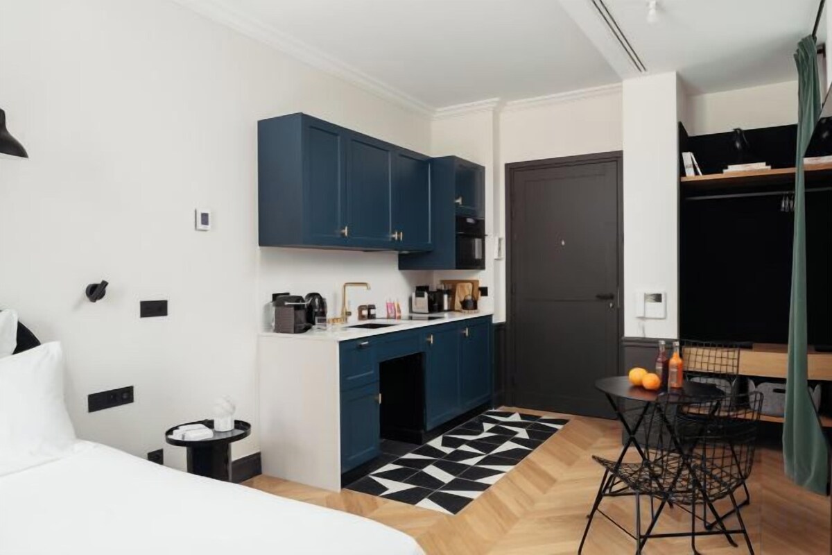A compact hotel room with a bed, a small kitchenette, a dining area, and a door leading to another room.