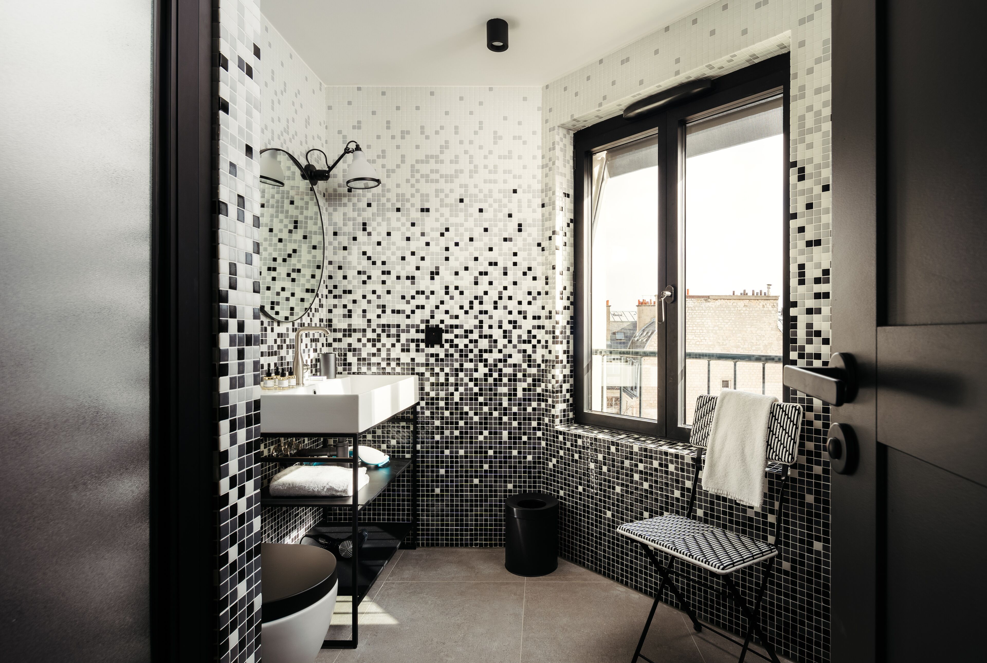 A compact bathroom with a tiled backsplash, a window with a view, a sink, a chair, and a trash bin.