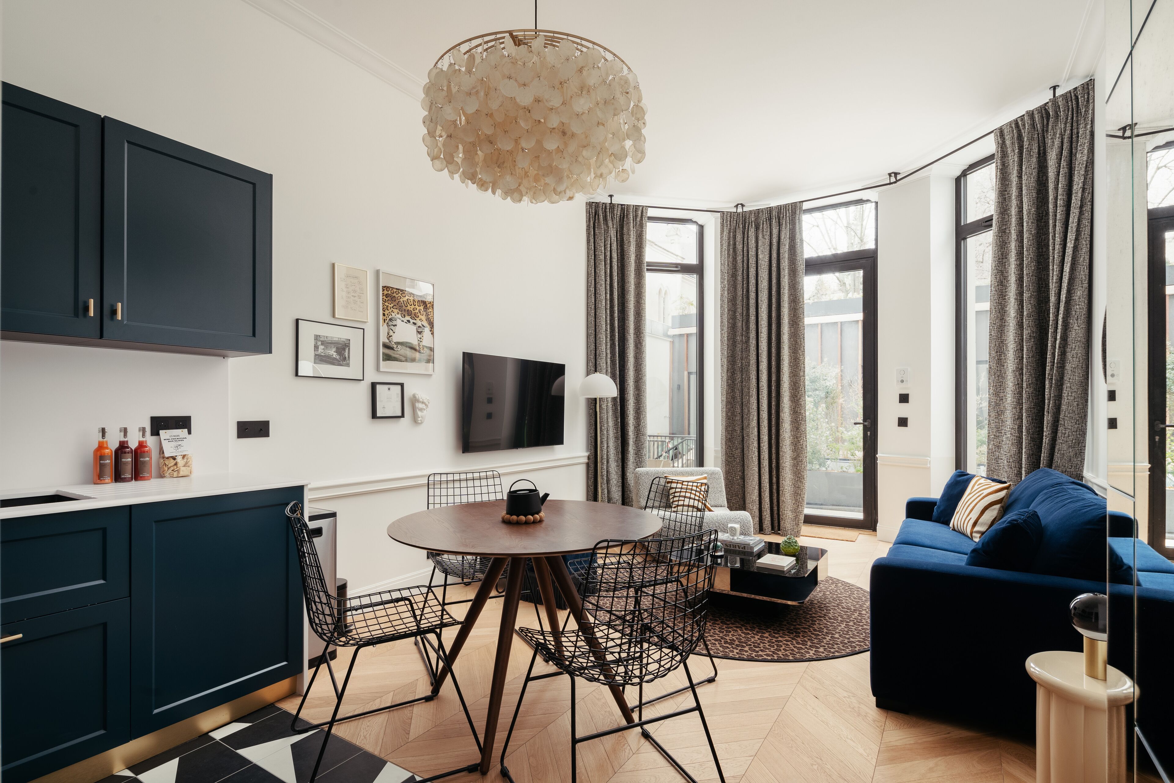 En moderne stue med en mørkeblå sofa, et rundt træbord og en sort metalstol. Der er et stort vindue med gardiner, et fladskærms-TV monteret på væggen og en dekorativ hængende lampe.