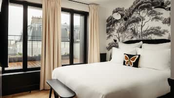 Habitación de hotel con una cama grande, un banco, vista a la ciudad a través de una ventana y un mural de árboles en la pared.