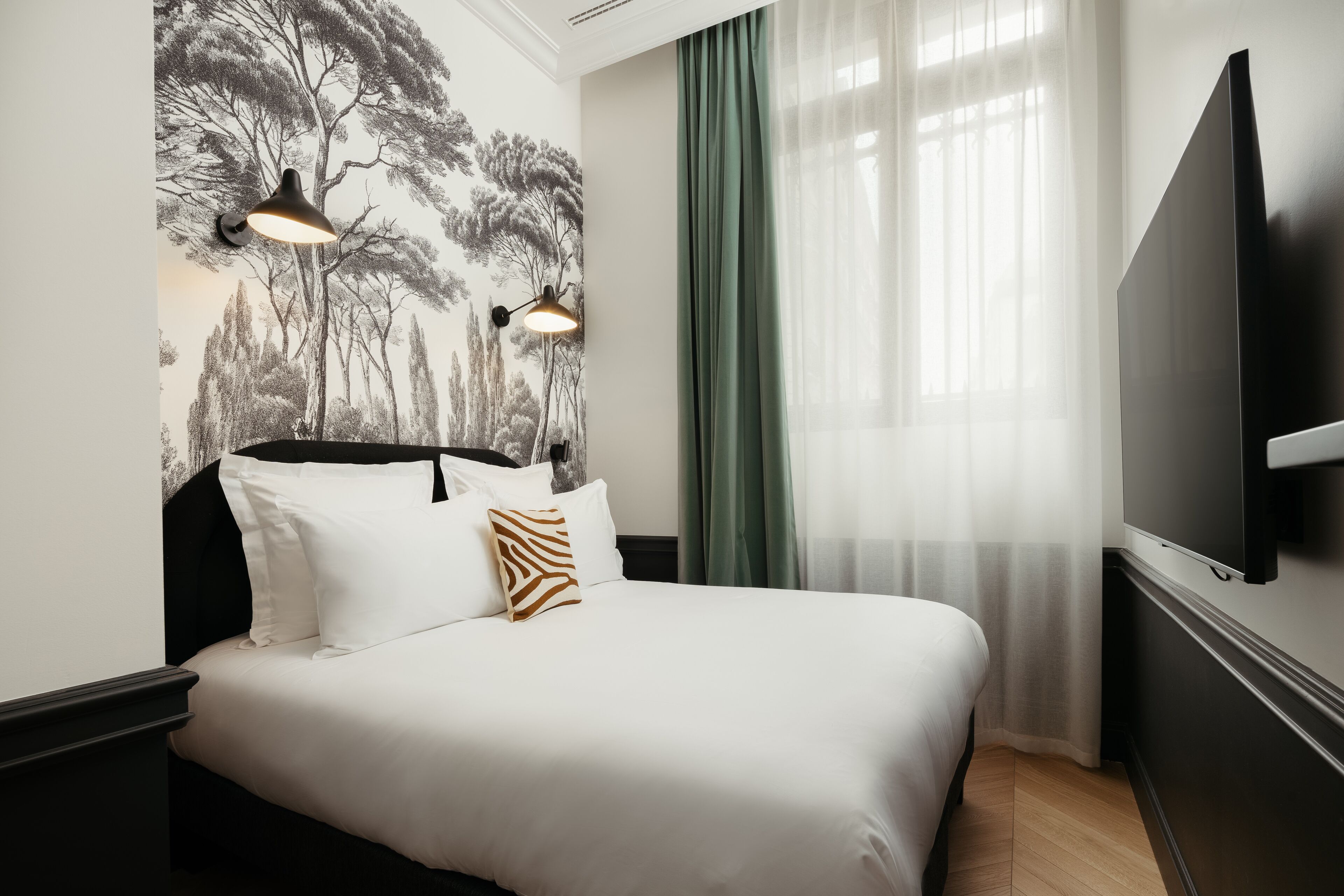 A hotel room with a bed, a wall-mounted TV, and a window with sheer curtains.
