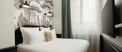 A hotel room with a bed, a wall-mounted TV, and a window with sheer curtains.